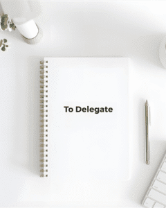"Minimalist white desk with a 'To Delegate' notebook, pen, keyboard, and plant, symbolizing the power to delegate.